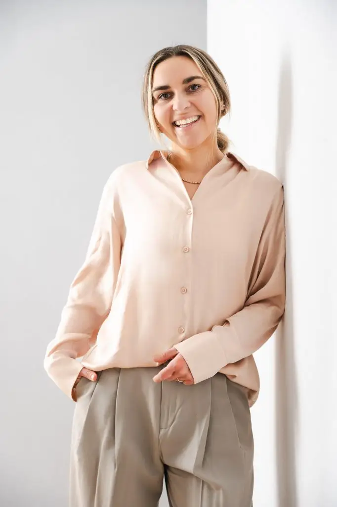 Portrait von Gründerin Eva Teufel. Sie lehnt an einer weißen Wand am rechten Bildrand. Sie trägt eine beige Businesshose und eine Rosa Bluse mit Kragen. Ihr blondes Haar trägt sie in einem tiefen, lockeren Pferdeschwanz. Zwei Strähnen rahmen ihr Gesicht ein. Ihre rechte Hand steckt in der Hosentasche. Eva Teufel lacht offen in die Kamera.