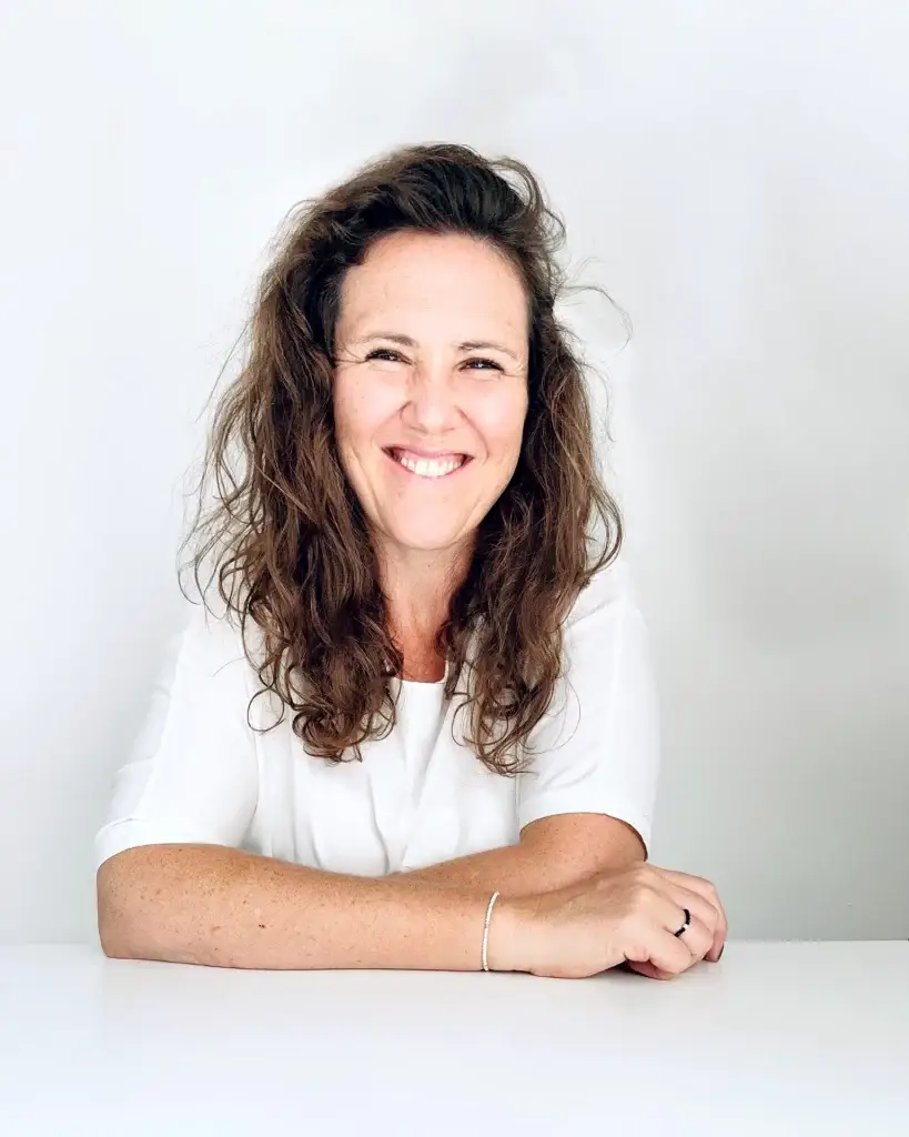 Portrait der Ansprechpartnerin Anita Klein für das Karrierecoaching beim gründungs.center. Sie lacht offen in die Kamera, ihre Haare sind offen. Sie trägt ein weißes Oberteil und sitzt vor einer weißen Wand und hat beide Arme auf dem weißen Tisch vor ihr liegen.