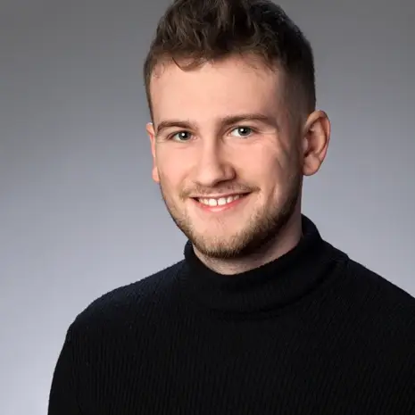 Portrait Teamassistenz Niklas Wolf: Ein blonder junger Mann lacht in die Kamera. Er trägt sein deckhaar länger, als seine Seiten, einen Bart und einen schwarzen Rollkragenpulli.