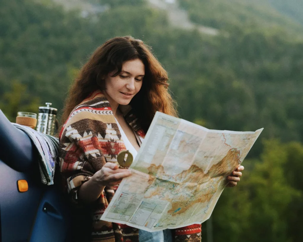 Symboldbild für "Womit als Frau selbstständig machen" für den Abschnitt "Menschen als Kompass". Eine Frau steht in der Natur, angelehnt an einem Auto und blickt auf die Landkarte in ihren Händen.