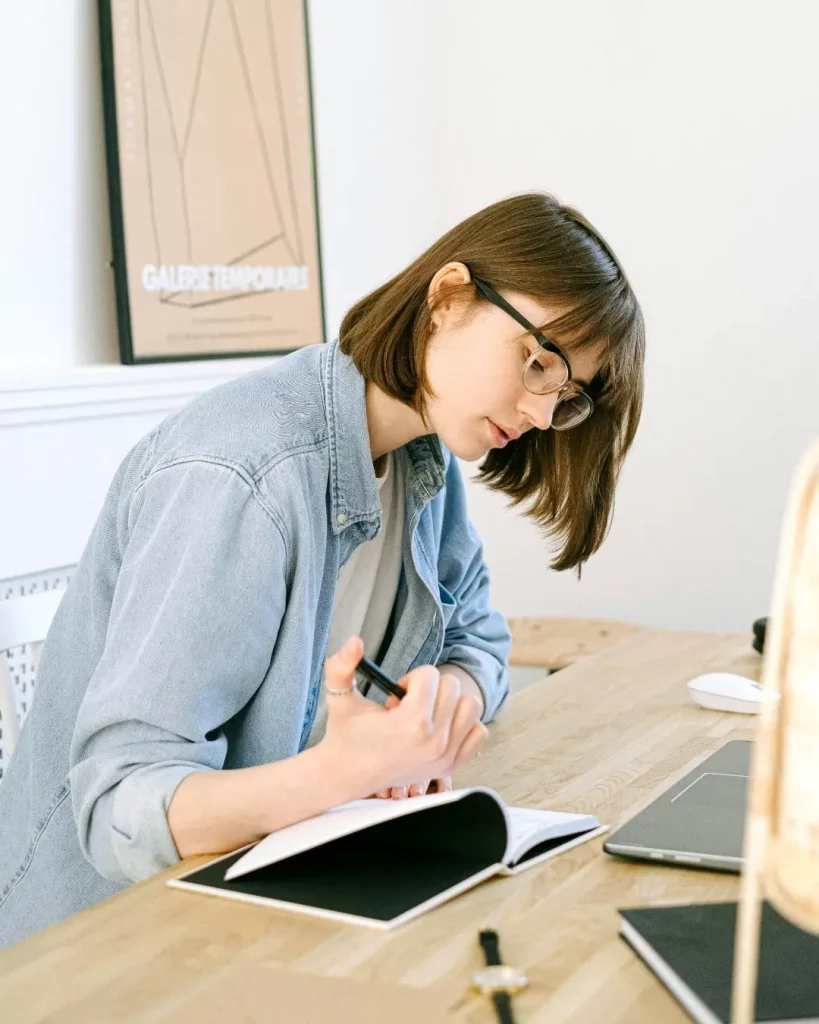 Symbolbild Fragen zum Bildungsgutschein: Eine junge, braunhaarige Frau sitzt übergebeugt an einem Schreibtisch mit Holztischplatte, an einem Laptop, in einem hellen Büro. Sie hält einen Stift in der Hand und trägt eine Brille. Es sieht aus, als würde sie etwas ausfüllen.