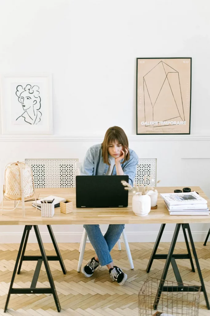 Symbolbild Fragen zum Bildungsgutschein: Eine junge, braunhaarige Frau sitzt übergebeugt hinter einem Schreibtisch mit Holztischplatte, an einem Laptop, in einem hellen Büro. Sie trägt eine Brille und schaut lustlos Richtung Laptop.