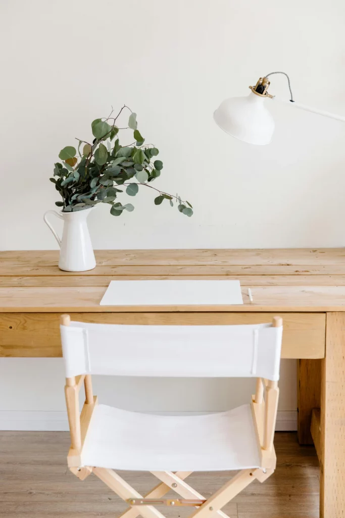 Symbolbild Fragen zum Bildungsgutschein: Ein helles Büro, mit einem Schreibtsich aus Holz, auf dem eine Vase mit Eukalyptuszweigen und eine Schreibtischlampe in weiß steht. Vor dem Schreibtisch steht ein Regiestuhl in weiß.