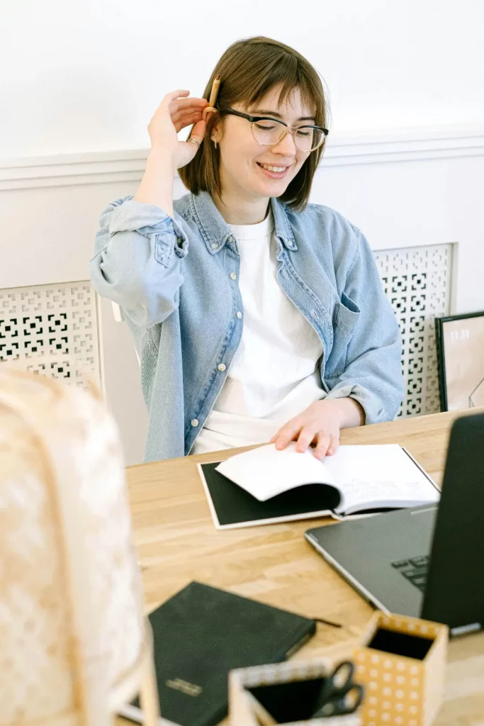 Symbolbild Fragen zum Bildungsgutschein: Eine junge, braunhaarige Frau sitzt hinter einem Schreibtisch mit Holztischplatte, an einem Laptop, in einem hellen Büro. Sie trägt eine Brille, streicht sich ihr Haar hinters Ohr und hält mit der linken Hand ein geöffnetes Notizbuch offen.