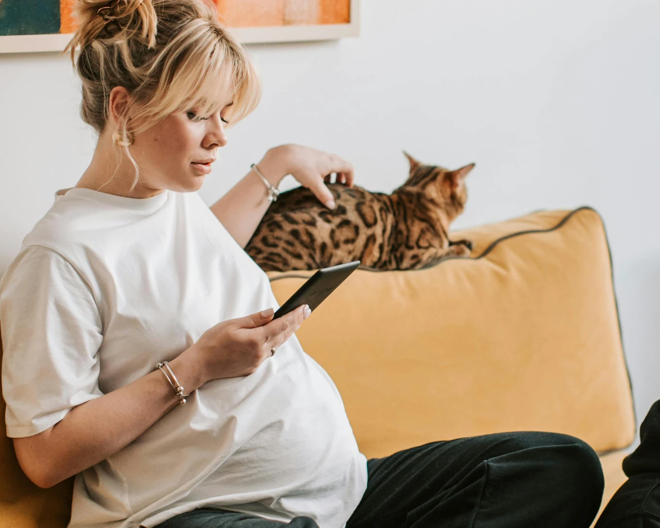 Sinn- und Coverbild: So gründest du in 5 Schritten als Mutter. Eine schwangere, blonde Frau, sitzt mit weiter Kleidung auf einem gelben Sofa. Sie blickt auf ein Handy und streichelt eine Katze.