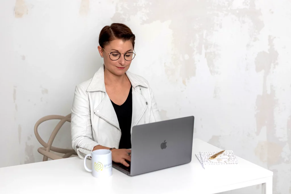 Sinnbild für Warum es unsere Ausbildung zum Gründungscoach gibt: Aline Pelzer sitzt in einem weiße Büro an einem Laptop und arbeitet daran.
